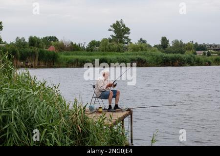 uomo anziano dai capelli grigi che pesca sul lago in natura, pazienza e concetto di rilassamento. pescatore con canna da pesca in natura pescatore seduto e pesce Foto Stock