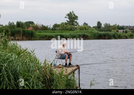 uomo anziano dai capelli grigi che pesca sul lago in natura, pazienza e concetto di rilassamento. pescatore con canna da pesca in natura pescatore seduto e pesce Foto Stock