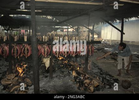 Un lavoratore sta mantenendo la distribuzione di buccia di cocco che brucia che è usato come combustibile per fumare le carni di tonno di skipjack ad un'industria domestica in Bitung, Sulawesi del nord, Indonesia. Secondo la FAO nel loro ultimo rapporto (2022), l'Indonesia è il terzo produttore mondiale di animali acquatici per la pesca e l'acquacoltura. Cinque paesi (Cina, India, Indonesia, Vietnam e Perù) sono responsabili di circa il 58% della produzione mondiale. Secondo la relazione, l'Indonesia è anche uno dei paesi in cui gli alimenti acquatici contribuiscono per la metà o più all'assunzione totale di proteine animali. Foto Stock