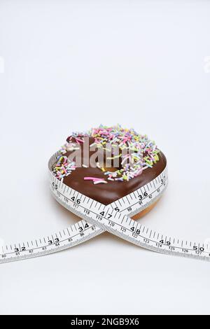 Un nastro di misurazione avvolto intorno a una ciambella ricoperta di cioccolato per illustrare quanto troppo cibo spazzatura può causare aumento di peso, su un concetto di sfondo bianco Foto Stock