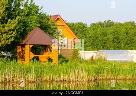 Giardini con cottage e gazebo per rilassarsi sulla riva di un piccolo lago in Siberia in estate tra l'erba. Foto Stock