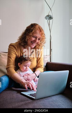 Donna sorridente mentre usa un computer portatile seduto sul divano con la figlia del bambino a casa. Foto Stock