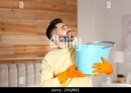 Giovane uomo che raccoglie l'acqua che fuoriesce dal soffitto a casa. È il momento di chiamare il servizio di riparazione del tetto Foto Stock