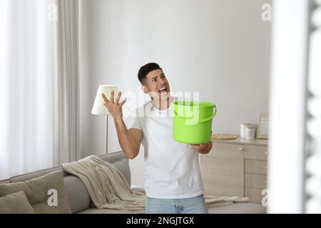 Emotivo uomo che raccoglie l'acqua che fuoriesce dal soffitto nel soggiorno. Tetto danneggiato Foto Stock