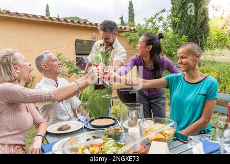 Gruppo di allegri amici di mezza età che brindano con un bel sorriso intorno al tavolo al patio della casa. Foto Stock
