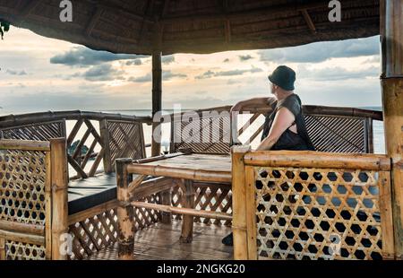 Seduti in un'area salotto ombreggiata in bamboo, accanto ad una spiaggia tropicale, guardando il sole d'oro, attraverso il Mare delle Filippine, sentendosi rilassati e tranquilli Foto Stock