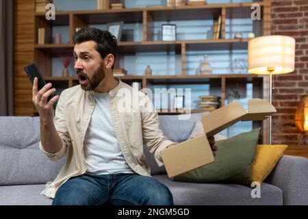 L'uomo ingannato arrabbiato a casa con telefono e pacco imballato ricevuto, discutendo al telefono parlare con il servizio di assistenza clienti del negozio online, la consegna del prodotto non è riuscita. Foto Stock