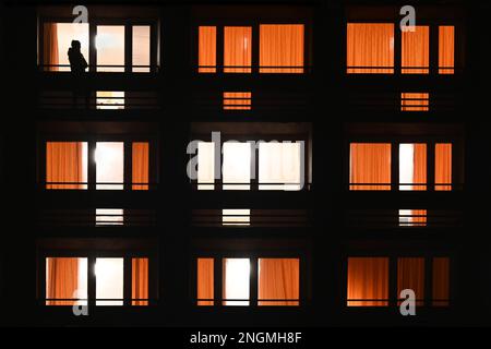 Finestre degli appartamenti illuminate di notte in un edificio urbano con silhouette di persona su un telefono o fumatori sul balcone Foto Stock