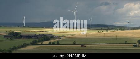 Paesaggio all'inizio dell'estate con sole che si infrange attraverso le nuvole scure ed evidenzia i campi con trattori in funzione e turbine eoliche a distanza ravvicinata Foto Stock