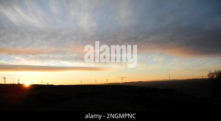 Paesaggio di tramontare il sole invernale all'orizzonte con la luce dorata riflessa nelle nuvole e il cielo con un gran numero di turbine eoliche in lontananza Foto Stock