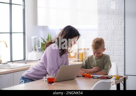 Lavora da casa e in famiglia, mamma e figlio utilizzano la tecnologia di accesso remoto per l'apprendimento e il lavoro, donna lavora sul notebook, bambino scrive nel libro di testo Foto Stock