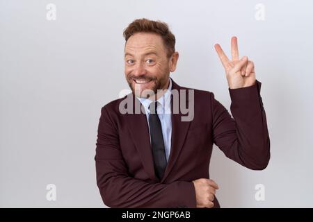 Uomo d'affari di mezza età con barba vestito e cravatta sorridente con faccia felice che si stringe alla macchina fotografica facendo segno di vittoria con le dita. numero due. Foto Stock