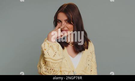 Ti sto guardando. Donna caucasica in cardigan puntando i suoi occhi e la macchina fotografica, mostra che sto guardando il gesto, spiando qualcuno. Giovane adulta bella ragazza isolata da sola su sfondo grigio parete studio Foto Stock