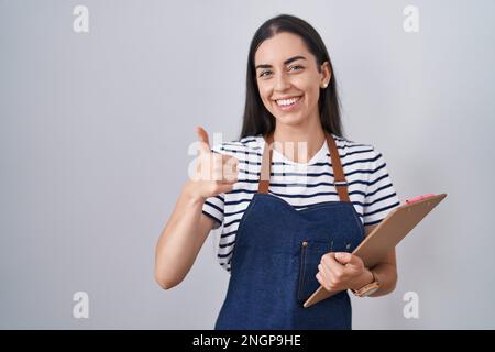 Giovane donna bruna indossando cameriera professionale grembiule e appunti sorridendo felice e positivo, pollice su facendo eccellente e segno di approvazione Foto Stock