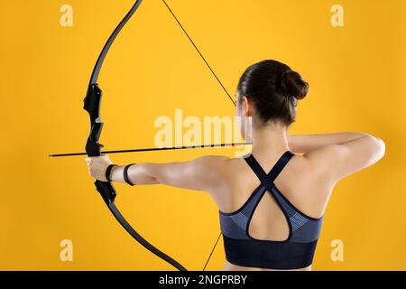 Donna con arco e freccia pratica tiro con l'arco su sfondo giallo, vista posteriore Foto Stock