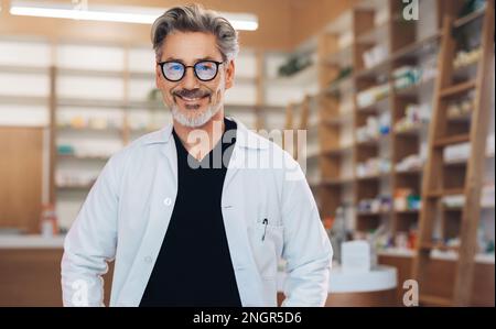 Ritratto di un farmacista maschile in piedi in una farmacia in un camice bianco da laboratorio. Uomo anziano che lavora in farmacia. Operatore sanitario maturo sorridente Foto Stock
