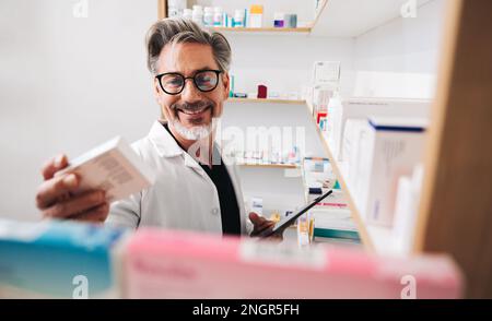 Il farmacista legge un'etichetta del farmaco e tiene una compressa in un chimico. Lavoratore di salute maschile maturo che riempie le prescrizioni online in una farmacia. Foto Stock