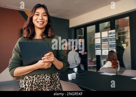 Allegra giovane donna d'affari che sorride alla fotocamera mentre tiene un tablet digitale. Felice giovane donna d'affari in piedi in una sala del consiglio con il suo collegagu Foto Stock