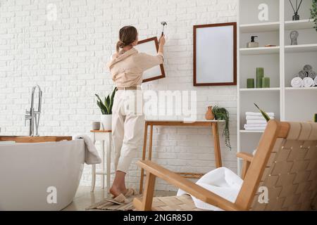 Giovane donna con martello appeso cornice vuota su muro di mattoni bianchi in bagno Foto Stock