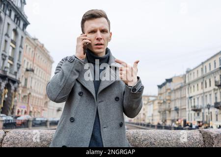 Ritratto giovane caucasico utilizzando il suo telefono cellulare in grigio cappotto si trova su una strada cittadina, un serio uomo d'affari occupato. Foto Stock