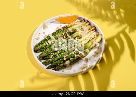 asparagi cotti in un piatto. asparagi fritti con olio d'oliva, conditi con salsa e cosparsi di noci, accanto ad una fetta di limone. Su un dorso giallo Foto Stock