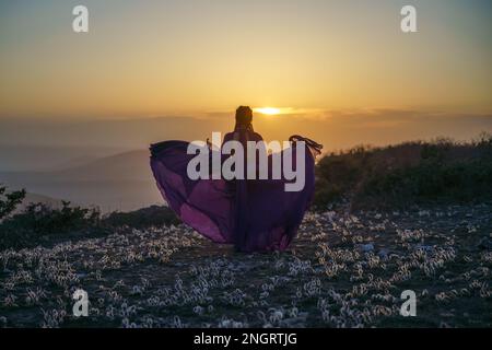 Tramonto viola abito donna montagne. Ascesa del mistico. tramonto sulle nuvole con una ragazza in un lungo vestito viola. Nel prato c'è un prato Foto Stock