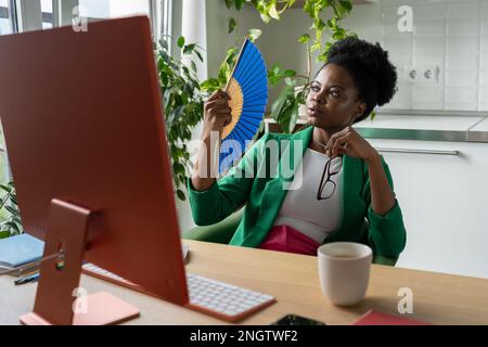 La donna d'affari africana usa il ventilatore della mano siede alla scrivania con il calcolatore in giorno di estate, soffre dal calore. Foto Stock