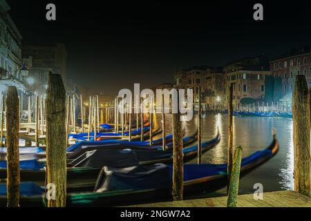 Venezia Italia Canal Grande vista notturna di edifici tradizionali e gondole vuote ormeggiate che oscillano accanto a pali di banchina in legno. Foto Stock