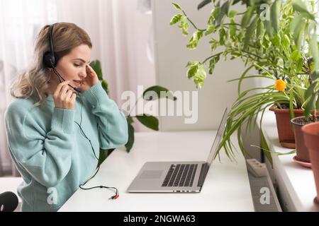 Vista laterale testa scattata sorridente signora freelancer gara mista indossando cuffie, comunicando con il cliente tramite videochiamata al computer. Piacevole millenario Foto Stock