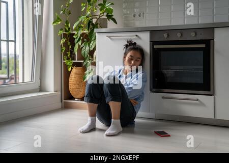 Frustrata ragazza asiatica triste seduta sul pavimento guardando il telefono cellulare che soffre di cattive relazioni. Foto Stock