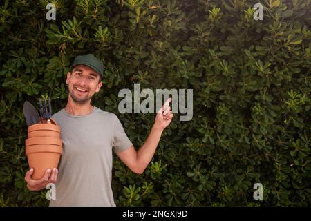 L'uomo giardiniere in tappo verde tiene pentole di terracotta con forniture di giardinaggio in mano. Punta il dito verso la recinzione sempreverde della matifolia di phillyrea. H Foto Stock