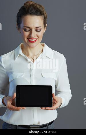 Donna sorridente piccola impresa di mezza età in blusa bianca che mostra uno schermo vuoto del Tablet PC isolato in grigio. Foto Stock