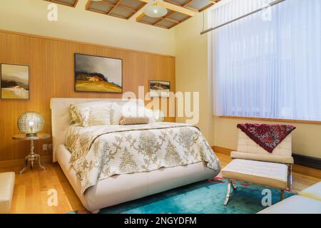 Letto queen size con testiera e struttura in pelle bianca, poltrona e ottomana nella camera da letto principale con pavimenti in legno di acero e ciliegio. Foto Stock