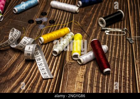 Una composizione di pezzi da cucire con varie bobine di filo colorato, bottoni, metro a nastro, forbici, un ago con filo filettato su un tavolo di legno Foto Stock
