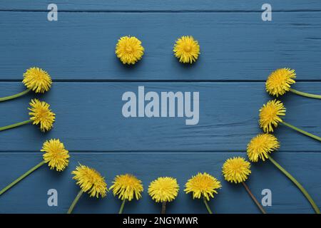 Faccia divertente fatta di bei dandelioni gialli su tavolo di legno blu, posa piatta Foto Stock