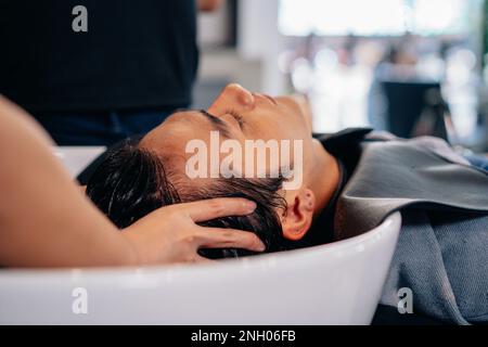 Bell'uomo asiatico che si rilassa con gli occhi chiusi con la testa nel lavandino mentre le mani di una donna parrucchiere lavano i capelli e premono i punti di pressione Foto Stock