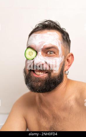 Uomo divertente con cetriolo e maschera di bellezza sul viso in bagno a casa Foto Stock