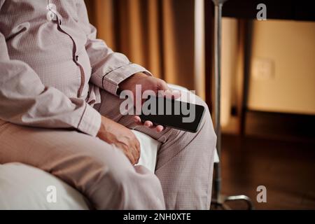 Immagine ritagliata di un uomo anziano seduto sul letto con lo smartphone in mano dopo aver ricevuto cattive notizie Foto Stock