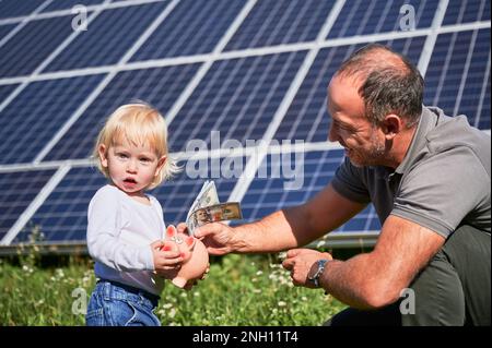 Padre e suo figlio piccolo mettere denaro in maialino banca per risparmiare denaro sullo sfondo di pannelli solari. Piccolo capretto interessato nel risparmio dei soldi per il futuro. Concetto di investimento in energia rinnovabile. Foto Stock