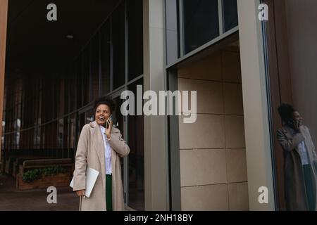 Una donna afroamericana d'affari con un portatile in mano cammina lungo l'edificio parlando al telefono Foto Stock