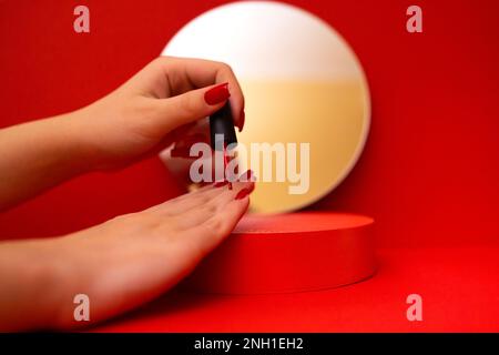 La giovane ragazza caucasica ha messo le mani su un podio rosso e le dipinge le unghie con pasta rossa. Uno specchio rotondo con glitter d'oro sullo sfondo. Primo piano. Foto Stock