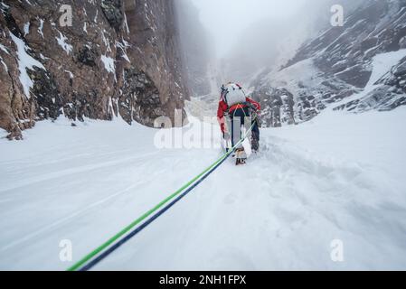 Arrampicatore di ghiaccio che arrampica sulla neve per arrivare alla parete rocciosa Foto Stock