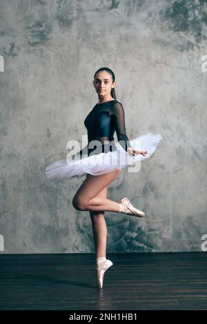 Corpo pieno di giovane ballerina sottile in tutu bianco e punta in piedi sui tipoes contro parete di scabro guardando la macchina fotografica Foto Stock