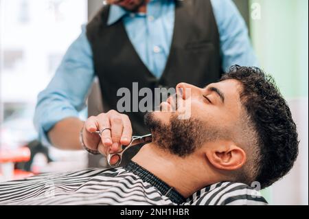 Crop barbiere anonimo utilizzando forbici, mentre il taglio della barba del cliente maschile in barbiere professionale Foto Stock