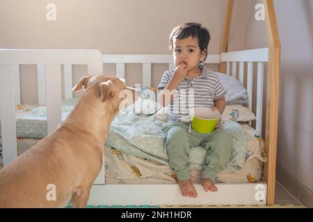 Piccolo ragazzo latino che mangia biscotti accompagnati dal suo cucciolo di razza mista Foto Stock