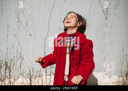 ragazza in cappotto rosso ridendo di fronte al muro di cemento con viti Foto Stock