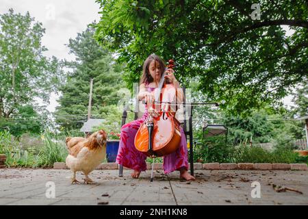 Una piccola ragazza a piedi nudi suona il violoncello per il suo pollo da compagnia in giardino Foto Stock