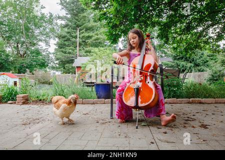 Un piccolo bambino felice suona il concerto di violoncello per il pollo dell'animale domestico all'esterno Foto Stock