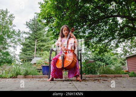 Un bambino piccolo gioca a violoncello a piedi nudi nel giardino sul patio Foto Stock