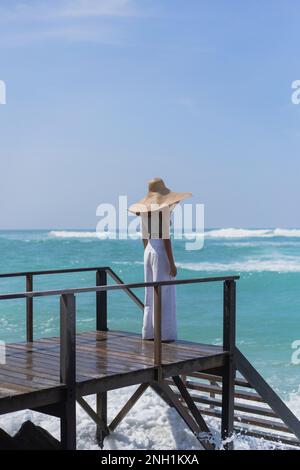 Giovane donna in un cappello di paglia sul molo, le onde dell'oceano schizzare. Foto Stock